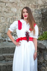Young woman in a white dress with a red sash and grape cluster accessory posing outdoors against a stone wall.