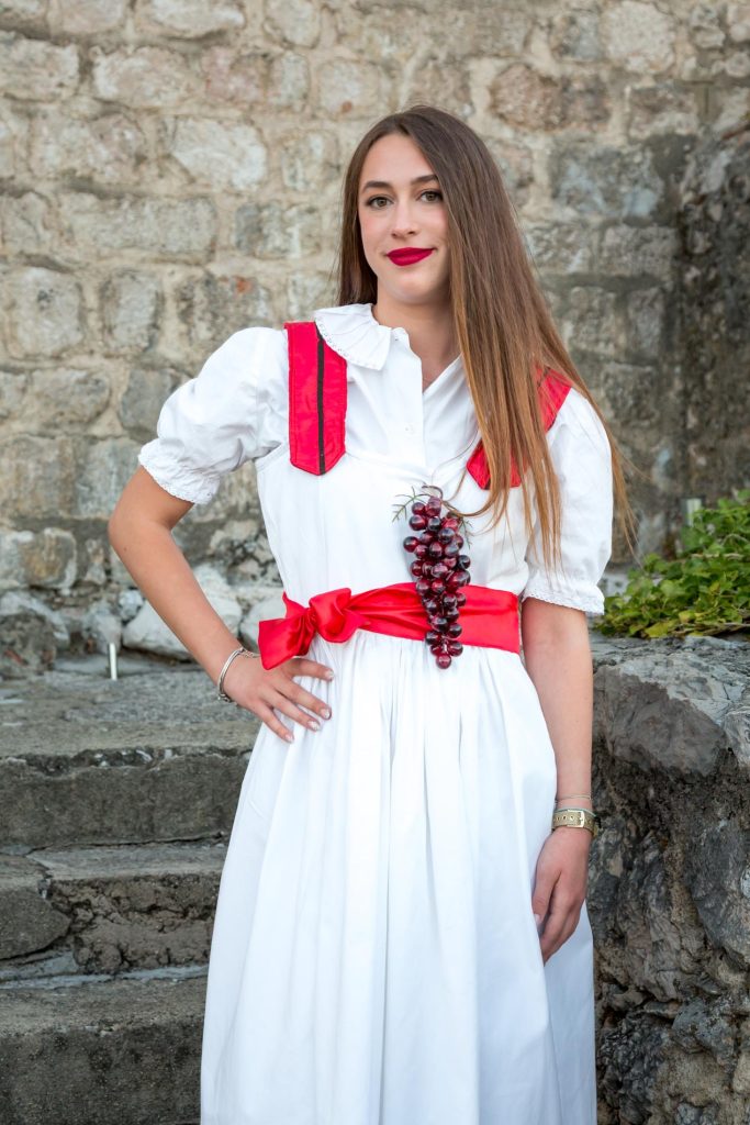 Young woman in a white dress with a red sash and grape cluster accessory posing outdoors against a stone wall.