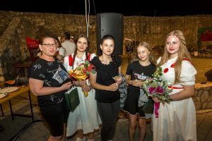Five people pose together at a nighttime outdoor event, holding bouquets and gift bags in front of a stone wall and speaker setup.