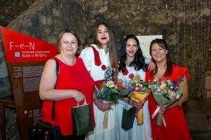 Four women pose together in a cave-like venue; two wear red dresses and two wear white gowns, each holding flowers and gift bags, smiling for the camera near a red informational sign.