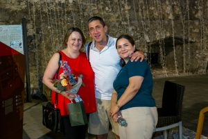 Three adults posing together in a stone-walled venue; woman in red holds a bouquet and gift bags with a man and another woman smiling beside her.