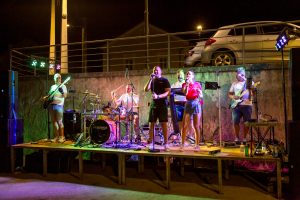 Band performs on a wooden outdoor stage at night, with drums, guitars, and microphones under colorful lights.