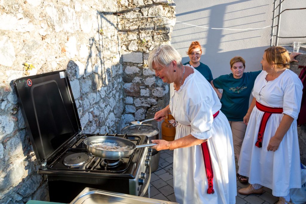 Older women in white dresses with red sashes cook on a grill outdoors beside a rough stone wall, with one woman stirring a pan over the flames.