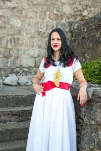 Woman in a white dress with a red sash and grape necklace, standing outdoors beside a stone wall, with dark hair and red lipstick.