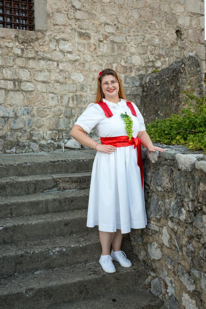 Smiling woman in a white dress with red sash and grape necklace stands on stone steps beside a rough stone wall, outdoors.