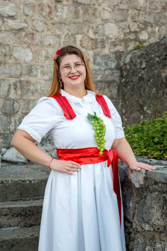 Smiling woman in a white folk dress with a red sash and grape necklace, posing against a stone wall outdoors.