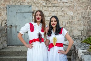 Two smiling women in white dresses with red sashes pose arm in arm in front of a stone wall, each wearing a grape necklace.