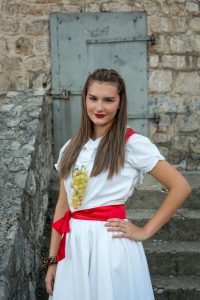 Young woman in a white dress with a red sash posing outdoors against a stone wall and metal door, wearing a grape cluster accessory and red lipstick.