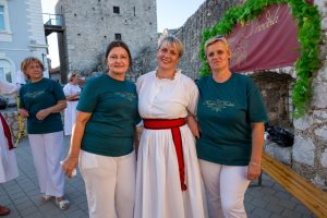 Three women posing outdoors in front of a stone wall; two wear teal tops with white pants, the center wears a white dress with a red sash and smiles at the camera.