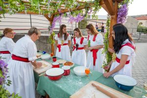 Group of women in white dresses with red sashes rolling dough at a decorated outdoor table under a flower-adorned pergola.
