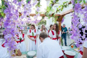 Group of people in white dresses with red sashes at an outdoor festival, framed by purple flowering vines on a wooden arch.