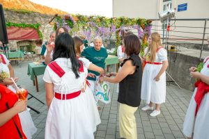Group of people at an outdoor festival, women in white dresses with red sashes and teal shirts under a purple flower arch. People share a moment near tables set for a celebration.