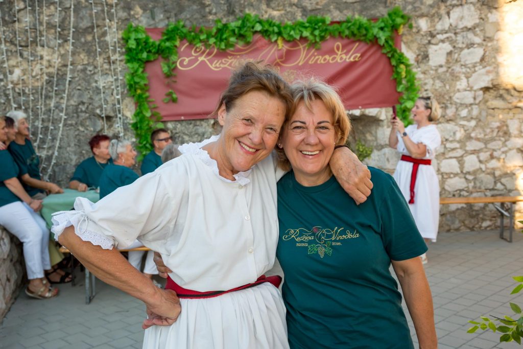 Two women smile and hug at an outdoor event, with a stone wall and greenery behind them.