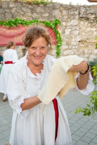 Smiling woman in a white traditional dress with a red belt holds up a light fabric outdoors in a stone courtyard festival setting.