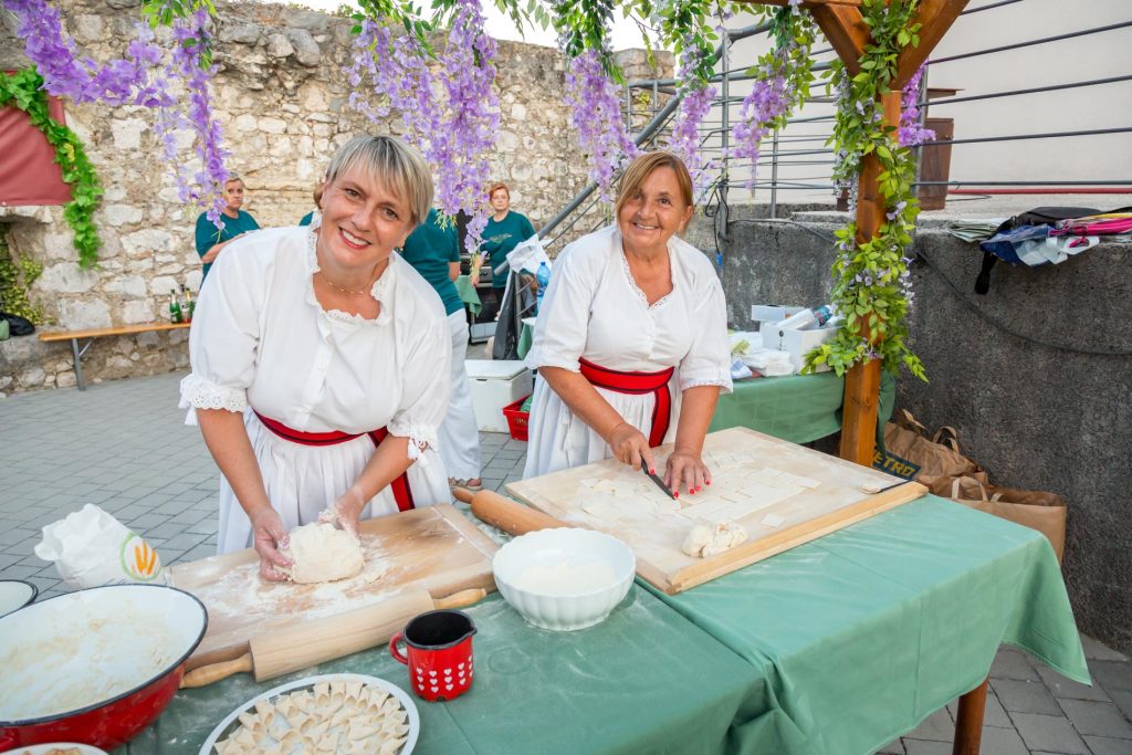Two women in white dresses with red belts roll and cut dough at an outdoor dumpling demo under hanging purple wisteria.