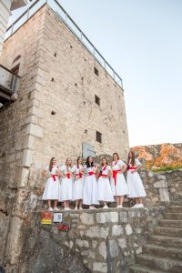 Group of women in white dresses with red sashes posing together on a stone platform in front of an old stone building against a clear blue sky