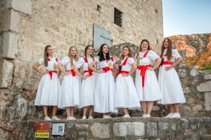 Group of women in white dresses with red sashes posing on a stone wall of an old fortress, hillside in background.