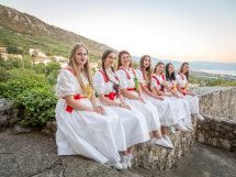Seven women in white dresses with red sashes sit on a stone wall overlooking a hillside village and sea.