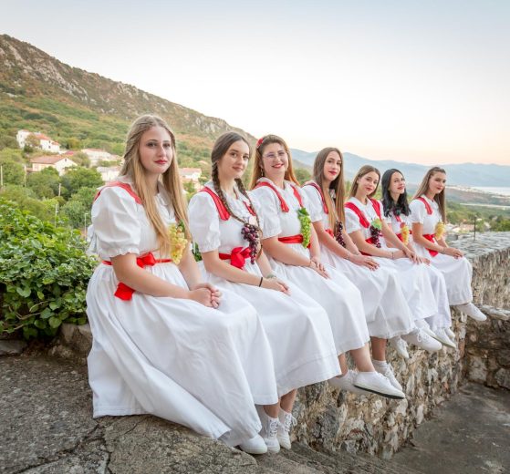 Seven women in white dresses with red sashes sit on a stone wall overlooking a hillside village and sea.