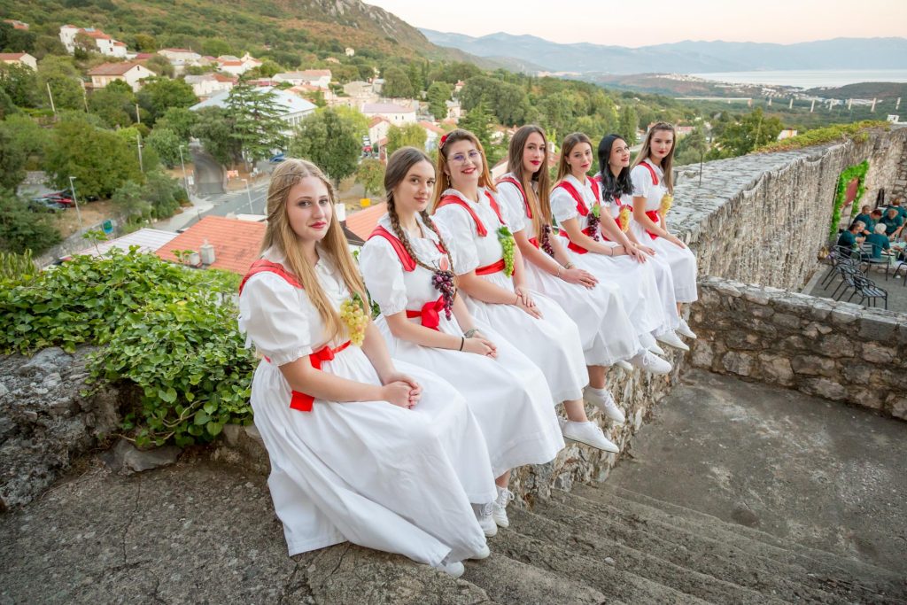 Group of young women in white dresses with red sashes sit on a stone ledge along a hillside, overlooking a village and distant mountains by the sea.