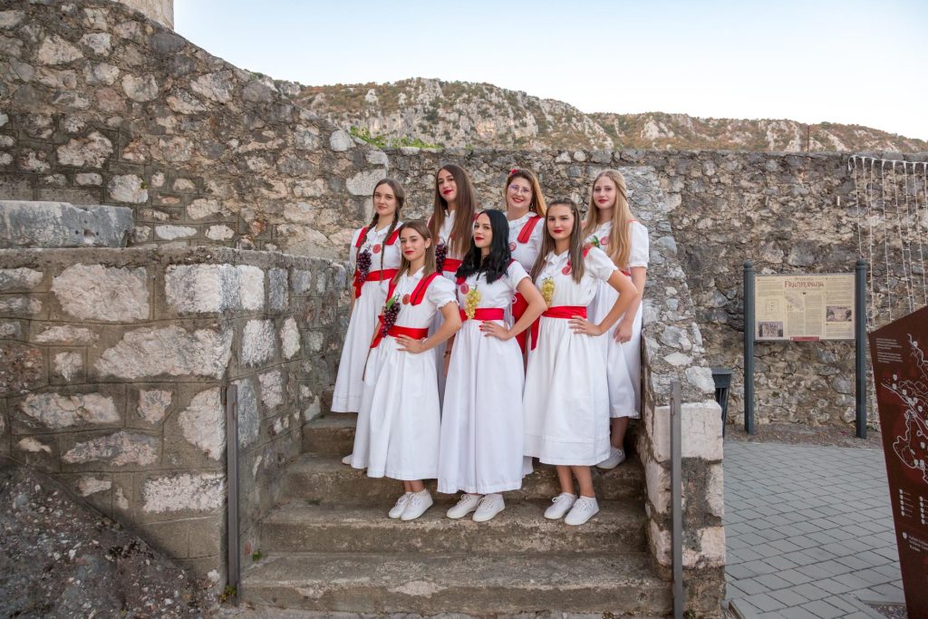 Eight women in white dresses with red sashes pose on stone steps at a historic fortress with a rocky hillside in the background.