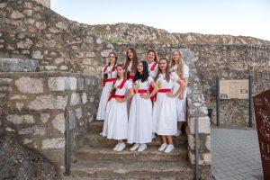 Eight women in white dresses with red sashes pose on stone steps at a historic fortress with a rocky hillside in the background.