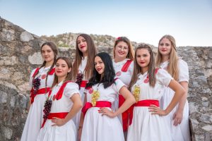 Group of young women in white dresses with red sashes posing together, each wearing grape clusters, in front of a stone wall outdoors.
