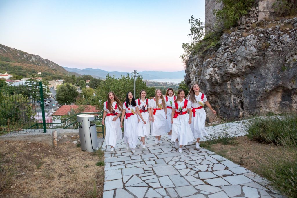 Group of nine women in white dresses with red sashes walking down a stone path with a rocky hillside and distant town and sea in the background.