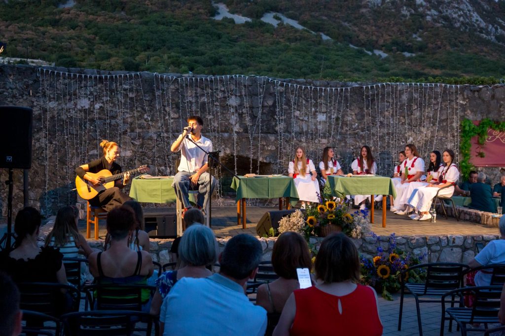Male singer with microphone and guitarist performing on an outdoor stage, with a row of girls in white traditional dresses at tables in front of a stone wall and string lights behind them.