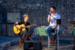 A woman playing a classical guitar while a man sings into a microphone outdoors against a stone wall with string lights behind them.