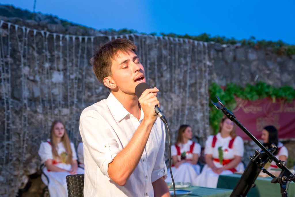 Male singer passionately performing into a handheld microphone outdoors, with a choir in white dresses behind him and string lights in the background.