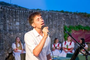Male singer passionately performing into a handheld microphone outdoors, with a choir in white dresses behind him and string lights in the background.