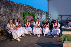 Five young women in white dresses with red sashes sit on chairs along a stone wall at an outdoor festival.