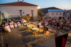 Outdoor cultural performance with dancers in white dresses seated on left, audience watching on terraces and chairs at dusk.