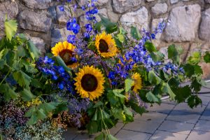 Bright bouquet of sunflowers, blue delphiniums, and greenery against a stone wall.