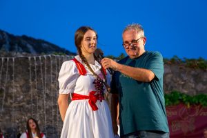 Young woman in a white folk dress with red sash and grape necklace being interviewed on a stage by a man holding a microphone, under a blue evening sky.