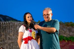 Two people on an outdoor stage: a woman in a white dress with grape decorations speaks into a microphone held by a smiling man in a teal shirt against a deep blue evening sky and hillside backdrop.