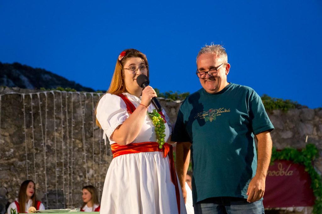 Woman in a white dress with a red sash speaks into a microphone on stage, standing beside a smiling man in a teal shirt.