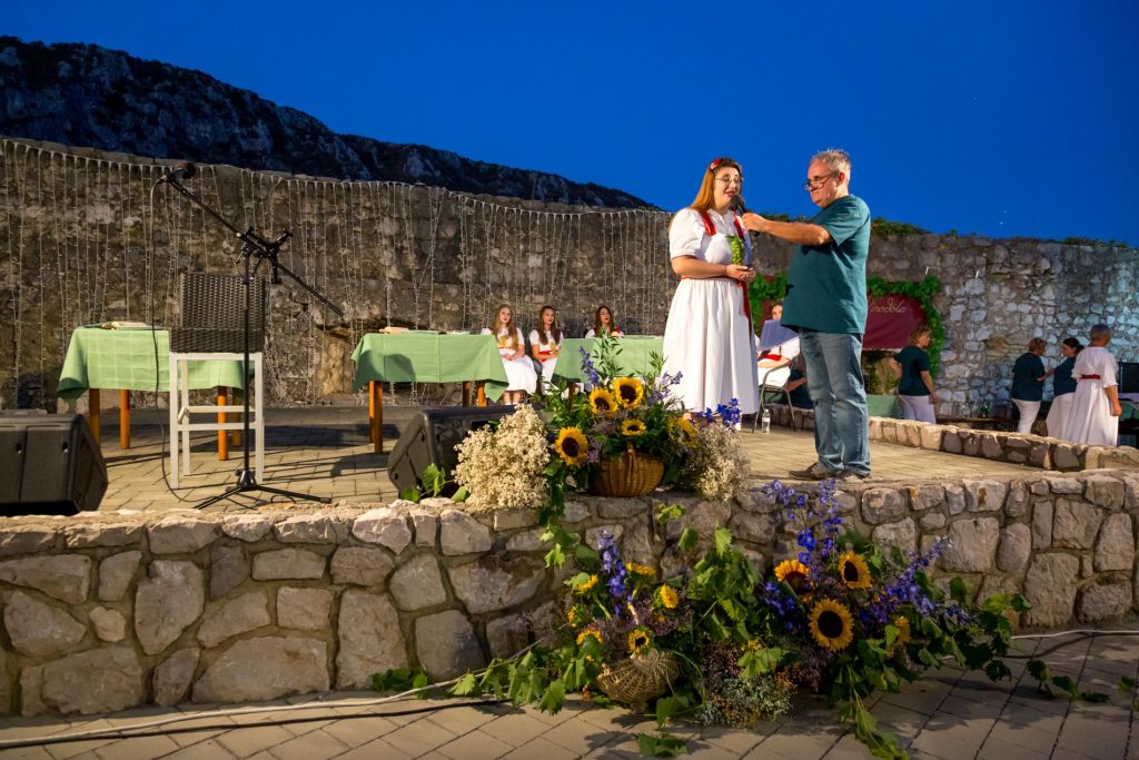 A woman in a white dress being interviewed on a stone outdoor stage, microphone held by a man in a teal shirt, with a large floral arrangement in the foreground and string lights along a stone wall behind them at dusk.