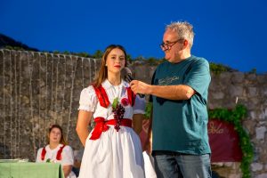Man with glasses holds a microphone to a woman in a white dress with red ribbons, grape accessory, on an outdoor stage at dusk.