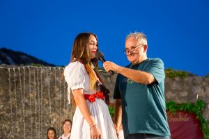 Man hands a microphone to a woman in a white dress with a red sash on an outdoor stage at night with a stone wall in the background.