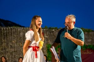 Young woman in a white dress with a red sash and grape necklace smiles on stage beside a man with a microphone at night.
