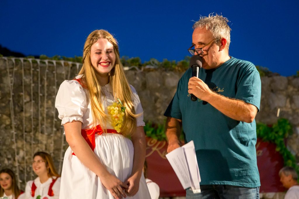 Young woman in a white dress with a grape garland smiles beside a man with a microphone at an outdoor event stage.