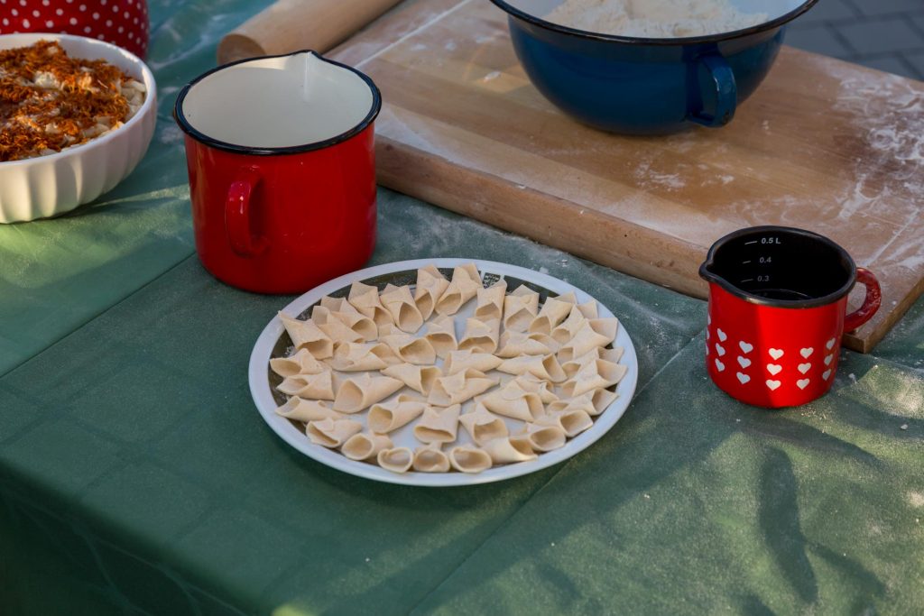 White plate filled with uncooked tortellini, beside red heart-pattern mug and a red mug on a green table, with a blue bowl and rolling pin nearby.