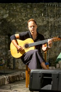 Woman seated outdoors, focused on playing a classical guitar during a performance, stone wall backdrop.