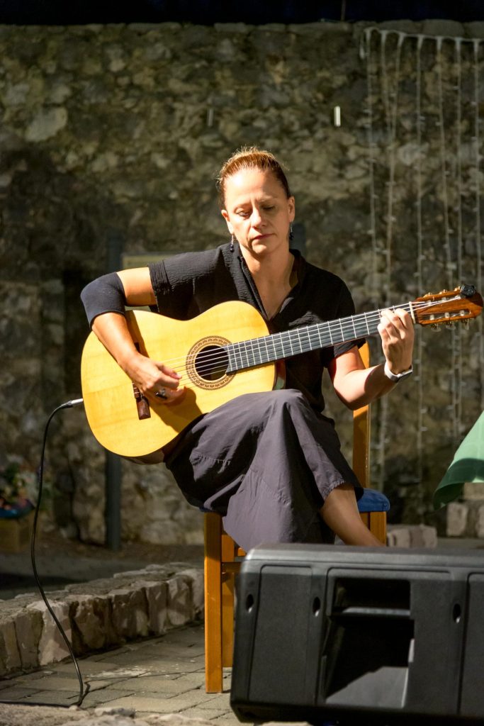 Woman seated outdoors, focused on playing a classical guitar during a performance, stone wall backdrop.
