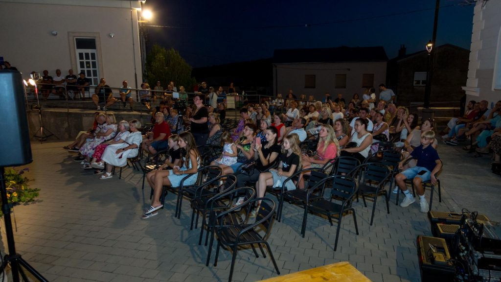 Audience seated on chairs in an outdoor night event, facing a stage on the left; a camera operator stands near center.