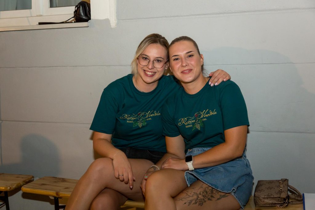 Two women sit close together on a wooden bench, smiling at the camera in matching teal shirts.