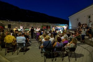 Outdoor evening concert with a singer at the microphone and a guitarist on a small stage, audience seated and watching.